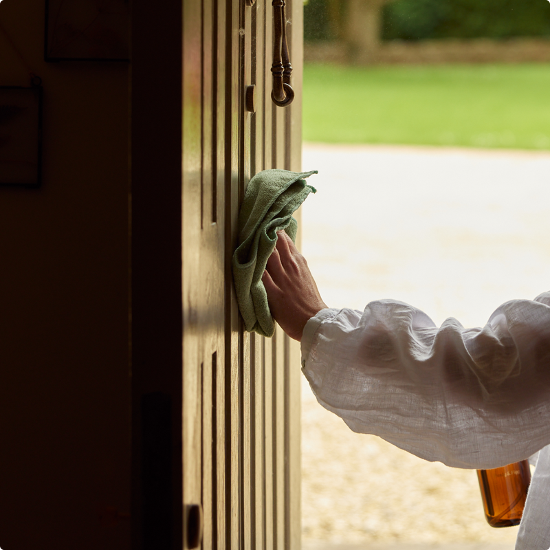 A woman in a white cotton shirt cleaning a wooden front door with a cloth and spray bottle